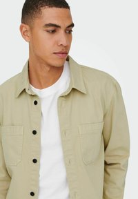 Hellbeige Button-Up-Jacke aus robustem Stoff, mit zwei Brusttaschen, schwarzen Knöpfen und einem weißen Rundhals-T-Shirt darunter.