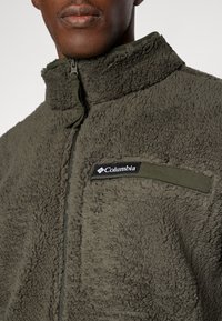 Olivgrüne Fleecejacke mit hohem Kragen, Frontreißverschluss und einer Brusttasche mit einem schwarzen Columbia-Logopatch. Weiche Textur.