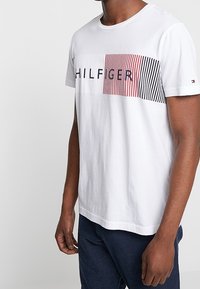 Bílý bavlněný tričko s grafickým designem horizontálních pruhů v růžové a černé barvě s nápisem "HILFIGER" v tučném písmu přes hrudník.