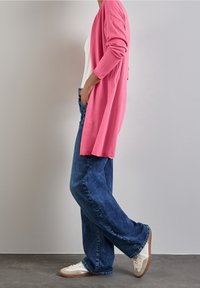 Dlouhý růžový cardigan s otevřeným předním dílem a žebrovanými manžetami, kombinovaný s volnými modrými džínami a bílými teniskami. Texturovaná tkanina, neformální styl.