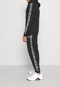 Svart hoodie och sweatpants med vitt Reebok-logotape längs sidorna, parat med vita löparskor. Enkel, sportig design.