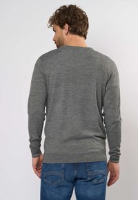 Mies, jolla on ruskeat hiukset, harmaa pitkähihaista neulepuseroa ja siniset farkut, seisoo selin yksinkertaista valkoista taustaa vasten.