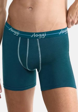 Mandlig overkrop iført tætsiddende turkise boxershorts med hvide syninger og mærket elastik i taljen, hånd justerer elastikken på højre side.