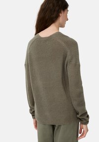 Strikket sweater i olivengrøn, med ribbet tekstur, drop-skuldrer og en rund bagkant. Båret med olivengrønne bukser.