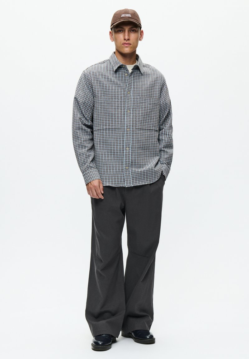 Young man wearing a brown cap, checkered button-up shirt, wide dark trousers, and black shoes, standing with one hand in pocket.