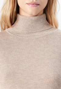 Blond kvinde iført en beige ribbet turtleneck sweater, der viser hals, læber og skuldre mod en hvid baggrund.