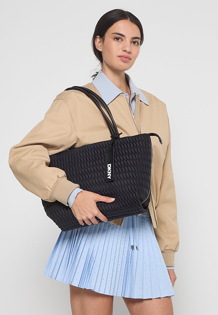 Junge Frau trägt eine beigefarbene Jacke und einen hellblauen plissierten Rock und hält eine große schwarze strukturierte DKNY-Umhängetasche.