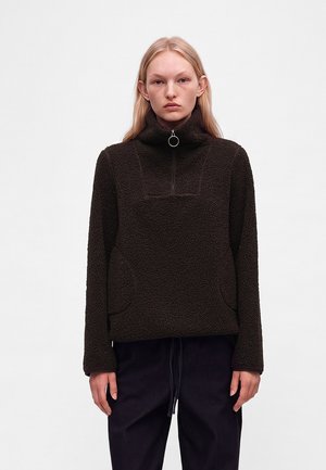 Ung person med glat blondt hår bærer en sort tekstureret fleece-pullover med høj krave og sølvringlynlås.