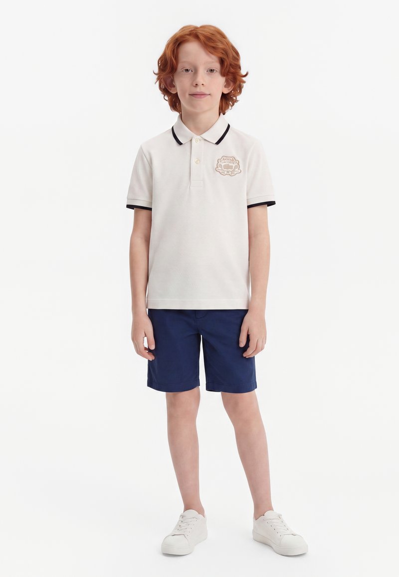 Enfant aux cheveux roux debout, portant un polo blanc avec un logo, un short bleu marine et des baskets blanches sur un fond blanc uni.