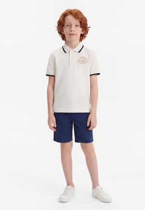 Enfant aux cheveux roux debout, portant un polo blanc avec un logo, un short bleu marine et des baskets blanches sur un fond blanc uni.