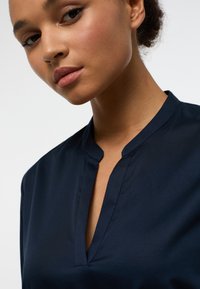 Blusa blu navy con una texture morbida, caratterizzata da un colletto alla cinese e un leggero scollo a V. Design pulito con dettagli di cucitura minimi.