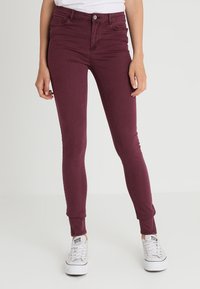 Vaqueros skinny burdeos en un material de denim elástico, con una cintura de altura media, bolsillos frontales y sutiles detalles de costura, combinados con zapatillas blancas.