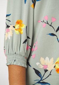 Blusa floreale con tessuto verde chiaro, motivi di fiori multicolori e maniche a sbuffo elastiche per un tocco di originalità e comfort.