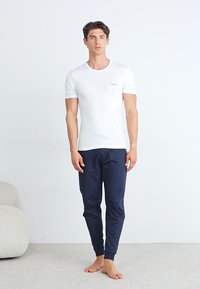 T-shirt blanche à manches courtes avec un logo discret, associée à un pantalon bleu marine ajusté. Texture de tissu lisse, design minimaliste.