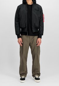 Giubbotto bomber nero con cappuccio, chiusura con zip, polsini a costine e tasca con etichetta rossa. Abbinato a pantaloni cargo oliva e sneaker nere.