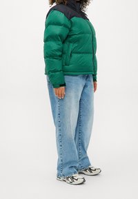 Zelená a černá péřová bunda s vysokým límcem, zipovým zapínáním, žebrovanými manžetami a dvěma bočními kapsami. V kombinaci s volnými modrými džíny a teniskami.