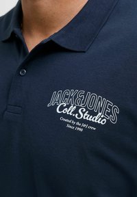 Marineblaues Poloshirt aus strukturiertem Stoff mit einem Knopfkragen. Es hat eine weiße gestickte Marke auf der Brust mit einem kleinen Logodetail.