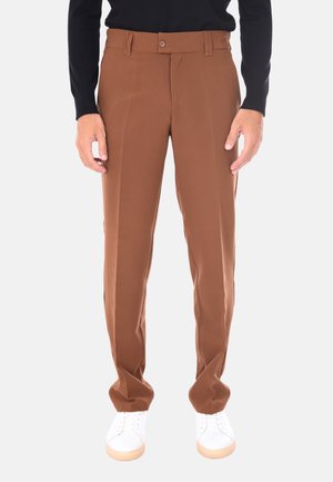 Uomo che indossa pantaloni marroni su misura, sneaker bianche e una maglietta nera a maniche lunghe, appoggiato a uno sfondo uniforme.