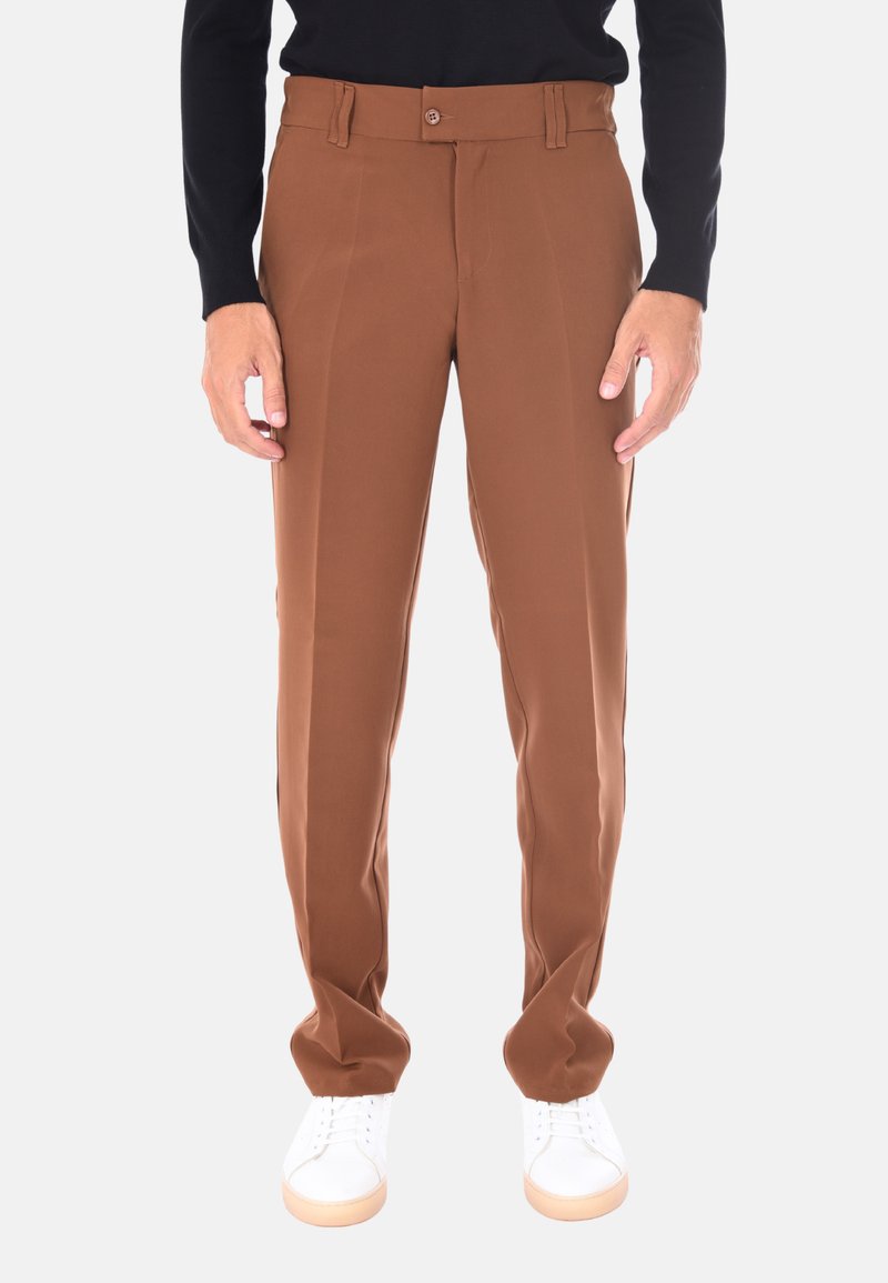 Uomo che indossa pantaloni marroni su misura, sneaker bianche e una maglietta nera a maniche lunghe, appoggiato a uno sfondo uniforme.