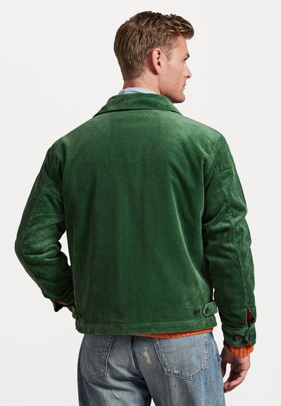 Giacca in corduroy verde con una finitura testurizzata, caratterizzata da un colletto, dettagli con bottoni e un design leggermente aderente, abbinata a jeans blu.