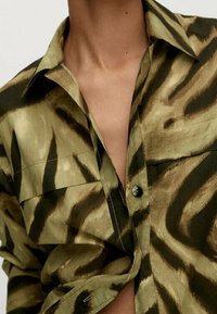 Short-sleeve shirt in a green and brown animal print. Features a collar, front pockets, and a single button closure. Smooth fabric texture.