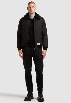 Schwarze Bomberjacke mit einem weichen Kragen, taillierter Passform und Reißverschluss, kombiniert mit schwarzen Hosen und klobigen schwarzen Stiefeln.