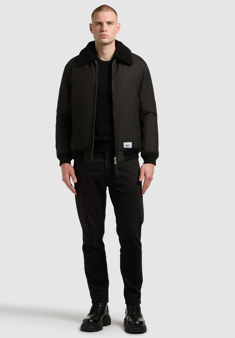 Schwarze Bomberjacke mit einem weichen Kragen, taillierter Passform und Reißverschluss, kombiniert mit schwarzen Hosen und klobigen schwarzen Stiefeln.