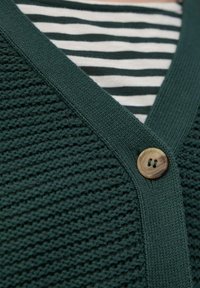 Dunkelgrüne Strickjacke mit V-Ausschnitt, strukturiertem Oberflächenmaterial und einem Holzknopf. Über einem weiß-grün gestreiften Hemd getragen.