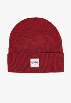 Red knit beanie made of soft fabric. Features a folded cuff with a white label displaying black brand logo. Textured ribbed design.