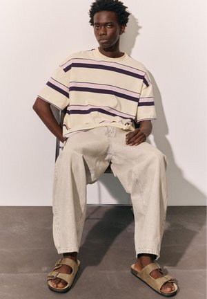 Giovane uomo seduto su una sedia che indossa pantaloni beige a gamba larga, una t-shirt a righe crema e viola e sandali beige con fibbie.