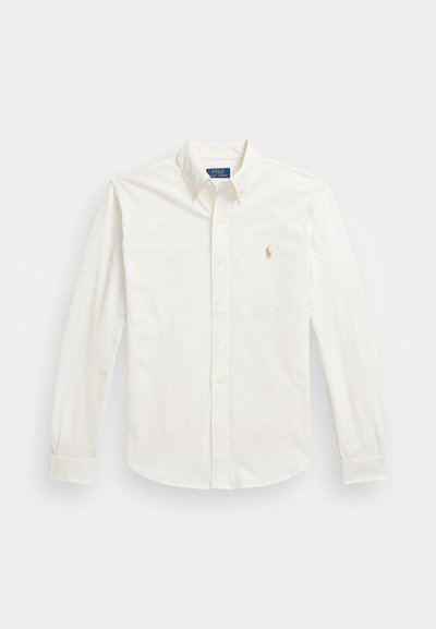 Hvit langermet skjorte i bomull med knapper, med spiss krage og en liten brodert logo på venstre bryst. Jevn tekstur.