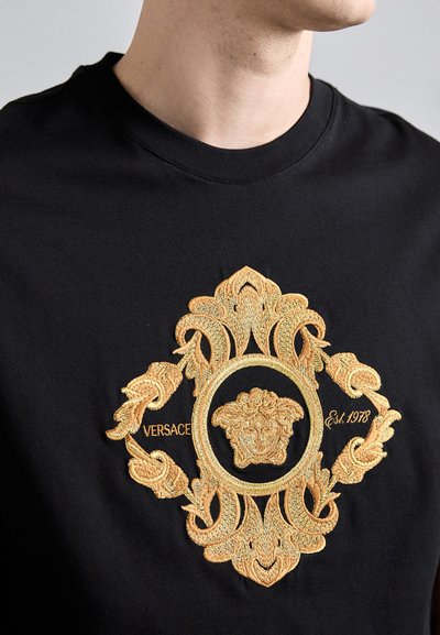 T-shirt en coton noir arborant un design brodé doré prominent avec des motifs élaborés et un médaillon central, étiqueté "Versace Est. 1978."