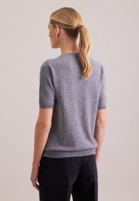 Grauer Pullover mit kurzen Ärmeln aus weichem Strick, rundem Ausschnitt, gerippten Bündchen und leicht taillierter Passform. Kombiniert mit dunklen, hoch taillierten Jeans.