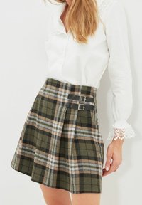 Vihreä ruutuhame, joka on valmistettu villakankaasta. Siinä on mustat metalliset soljet vyötäröllä ja pleatattu muotoilu. Yhdistä valkoisen paidan kanssa.