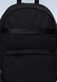 Svart ryggsäck i texturerad tyg med ett kviltat mönster. Framtida dragkedjefack med en märkesrem överst.