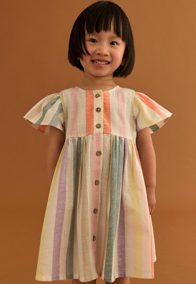 Junges Kind lächelt, trägt ein kurzärmeliges, geknöpftes Kleid mit vertikalen Pastellstreifen vor einem schlichten braunen Hintergrund.