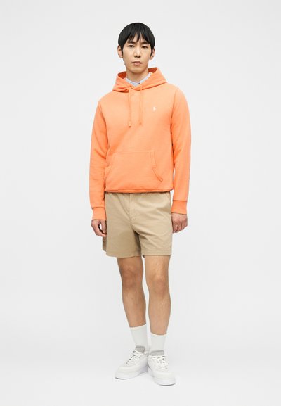 Νεαρός άνδρας με κοραλλί φούτερ με κουκούλα, μπεζ σορτς, λευκές κάλτσες και λευκά αθλητικά παπούτσια, στεκόμενος μπροστά σε απλό λευκό φόντο.