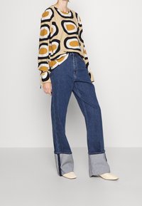 Beiget långärmad skjorta med svarta och senapsgula abstrakta mönster, tillsammans med blå vida jeans med uppvikta ben i ljusare denim.