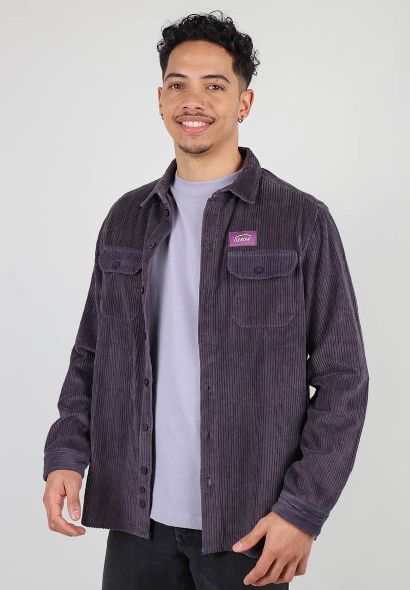Chemise en velours côtelé violet avec deux poches poitrine, fermeture à boutons, et un patch logo cousu. Portée par-dessus un T-shirt gris clair, coupe droite.