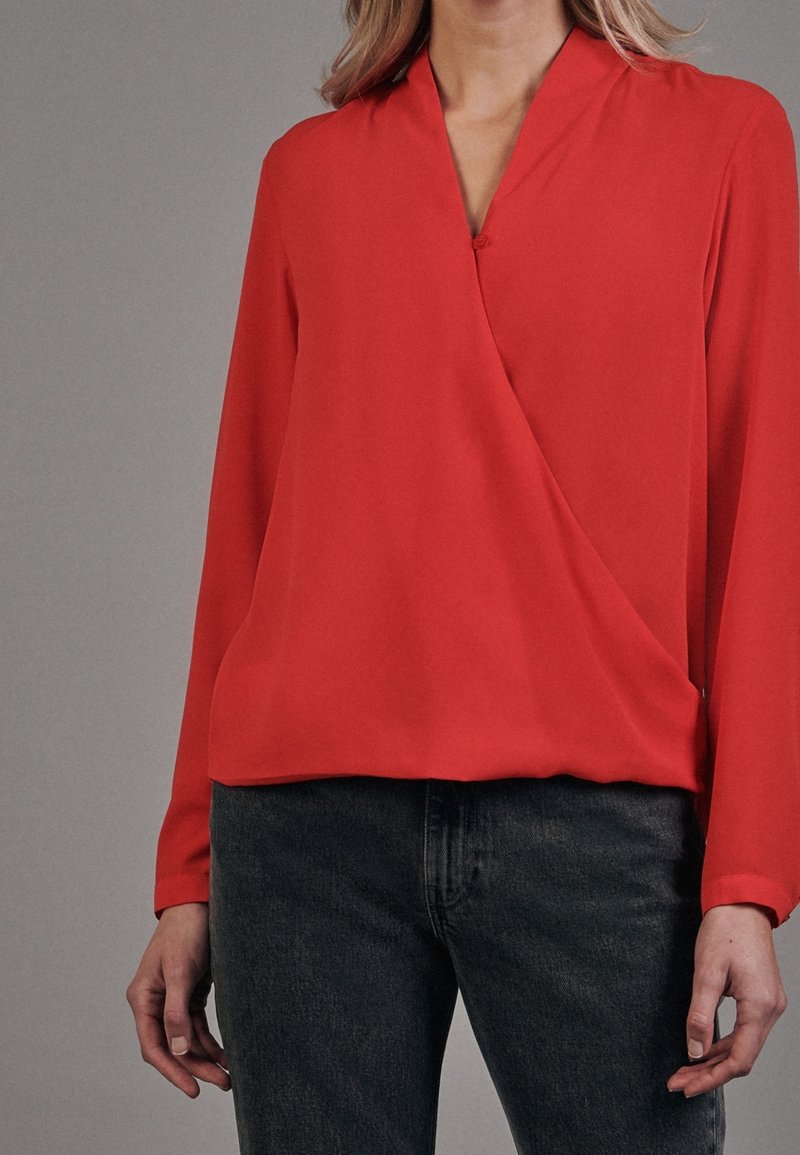 Blouse rouge à manches longues avec un col en V et un design portefeuille, fabriquée en tissu lisse. Associée à un jean foncé et ajusté. Design minimaliste, sans motifs.