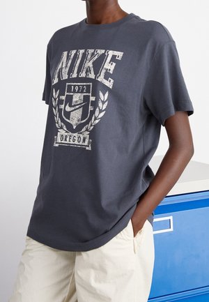 Person iført en mørkegrå Nike t-shirt med hvidt logo og tekst, kombineret med lyse beige bukser, stående nær et blåt skab.