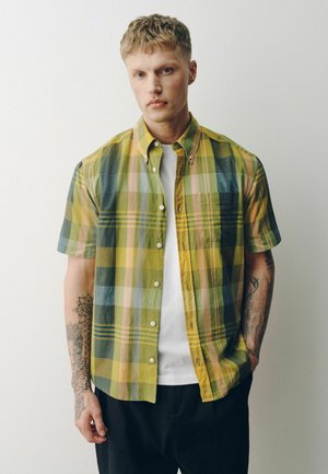 Young man with short hair, wearing a yellow and green plaid short-sleeve shirt over a white t-shirt, standing against a white background.