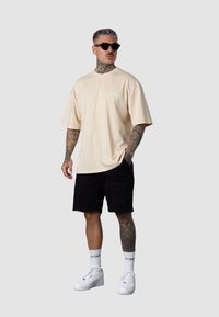 Beige übergroßes T-Shirt, schwarze Shorts, weiße Sneaker und Sonnenbrille. Tätowierungen an den Armen sichtbar. Schlichtes Design, entspannter Schnitt.