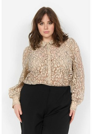 Lace blouse in beige with floral patterns, collared design, and puffed sleeves. Paired with black trousers, featuring a classic button-up front.