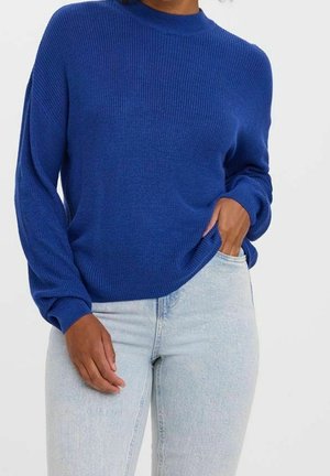 Femme portant un pull bleu à manches longues et un jean bleu clair, une main dans la poche et l'autre détendue le long du corps.