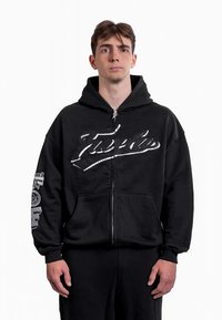 Sudadera con cremallera negra y un bolsillo frontal. Presenta la palabra "Truth" en blanco en el pecho y diseños gráficos en la manga izquierda. Tejido suave.