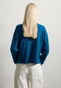 Suéter corto de color teal con un acabado texturizado, usado sobre pantalones crema. Cabello largo y lacio de color rubio visible en la parte de atrás. Fondo simple.