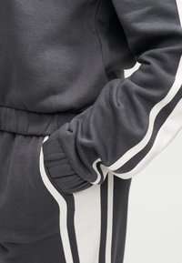 Γκρι φόρμα με λευκές πλαϊνές ρίγες. Το υλικό είναι απαλό, με ριμπ μανσέτες και ελα στική μέση για άνετη εφαρμογή.