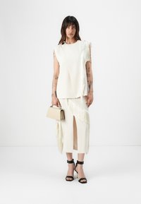 Cream-colored two-piece outfit with fringes, featuring a sleeveless top and a skirt with a thigh-high slit. Black strappy heels and small bag.