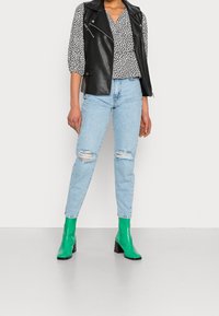 Gilet di pelle nera sopra una camicetta bianca con pois neri, jeans strappati azzurri chiari e stivaletti verdi con tacco a blocco.